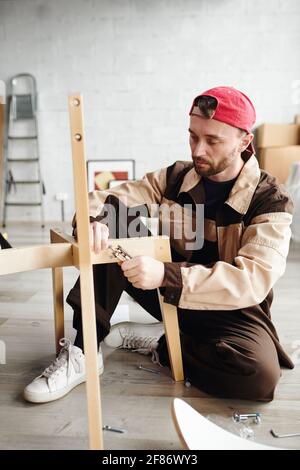 Serious junge Möbel Assemblage Spezialist in Arbeitskleidung sitzen auf der Boden und Befestigungsteile des Stuhls während der Montage Neue Wohnung oder Studio Stockfoto
