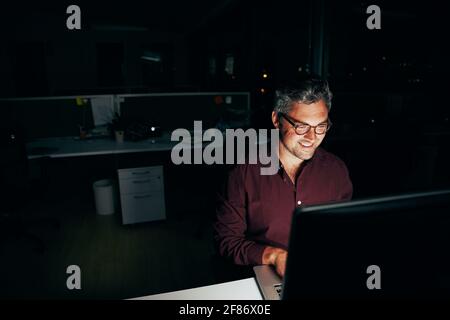 Porträt eines lächelnden Geschäftsmannes, der an einem Computer arbeitet Sein Schreibtisch bis spät in die Nacht Stockfoto