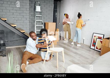 Der junge Afrikaner und sein süßer kleiner Sohn bauen Holz zusammen Stuhl, während er auf dem Boden sitzt, gegen zwei Frauen, die malen Wand in weißer Farbe Stockfoto