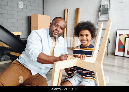 Der junge Afrikaner und sein fröhlicher kleiner Sohn bauen Holz zusammen Stuhl, während er in der neuen Wohnung auf dem Boden sitzt Oder Haus Stockfoto