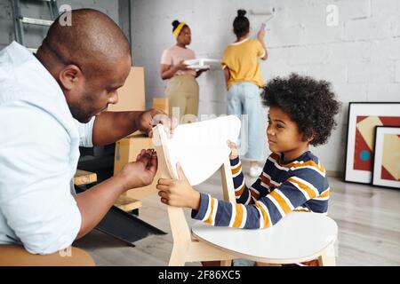 Netter kleiner Junge, der seinem Vater dabei hilft, einen Holzstuhl zusammenzustellen Gegen Mutter und Schwester Malerei Wand des Wohnzimmers in weiß Farbe Stockfoto