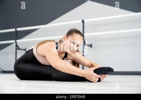 Anmutige, flexible, attraktive Frau, die sich auf dem Boden streckt, die Kamera anschaut und lächelt. Erwachsene Sportlerin, die das Gesicht auf die Knie legt und vor dem Wettkampf im Tanzsaal trainiert. Konzept der Gymnastik. Stockfoto