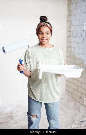 Glückliches Teenager-Mädchen mit Paintroller und weißem Behälter mit Farbe An zwei Wänden vor der Kamera gegen eine Ecke stehen Des Wohnzimmers Stockfoto