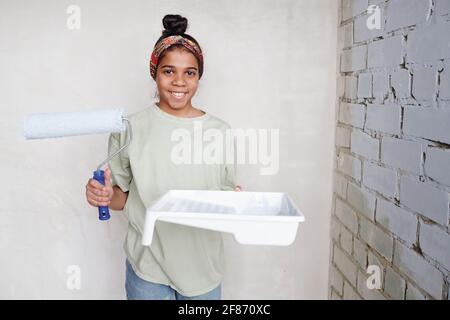 Happy niedlichen Teenager-Mädchen mit Paintroller und weißen quadratischen Kunststoff Behälter mit Farbe, die in der Ecke an zwei Wänden steht Des Wohnzimmers Stockfoto