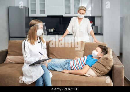 Kompetenter Kinderarzt in Schutzmaske und Handschuhe schreiben auf Zwischenablage während der Untersuchung von Teenager. Kranker Junge liegt auf der Couch und Mutter steht in der Nähe zu Hause. Stockfoto