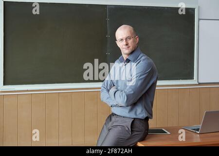 Kahlköpfiger Universitätsprofessor in intelligenter Casualwear, der auf dem Schreibtisch sitzt Gegen die Tafel, während die Arme an der Brust überquert und betrachtet werden Sie Stockfoto