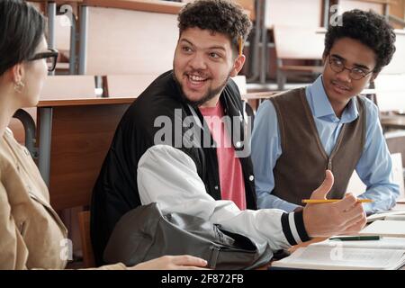 Glücklicher Kerl mit Bleistift, der einen seiner Gruppenmitglieder ansieht Während der Diskussion des Projektthemas oder der Passage aus dem Buch bei Unterricht im Hörsaal Stockfoto