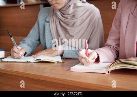 Hände von zwei jungen Studentinnen mit Stiften, die Notizen machen In Copybooks bei der Vorlesung, während sie in Reihe an Lond sitzen Holzschreibtisch im Auditorium Stockfoto