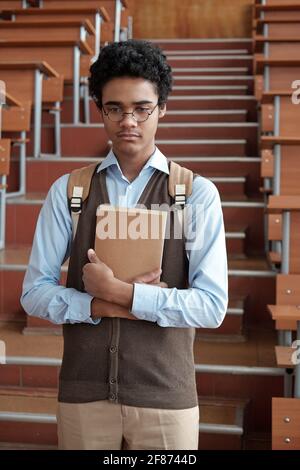 Nachdenklich oder beleidigt interkulturellen männlichen Studenten in Smart Casualwear und Brillen, die das Buch halten, während sie im Auditorium gegen die Treppe stehen Stockfoto