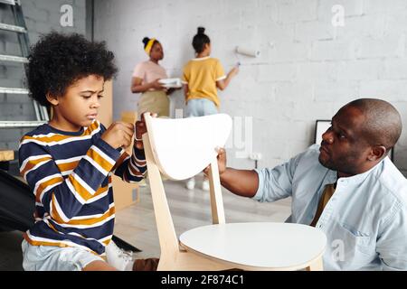 Netter kleiner Junge, der seinem Vater dabei hilft, einen Stuhl zusammenzustellen Fixing seinen Rücken gegen Mutter und Schwester Malerei Wand von Wohnzimmer in weißer Farbe Stockfoto