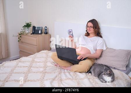 Dollar in den Händen von Schwangeren auf dem Bett im Home Office, Telearbeit und Geld Stockfoto