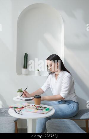 Ziemlich junge kreative Kunststudentin, die in der Cafeteria am Tisch sitzt Und zeichnen Bild mit Buntstiften in Skizzenbuch Stockfoto