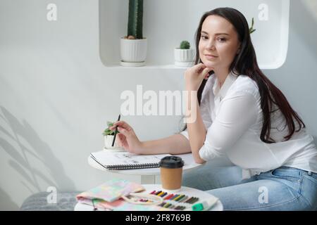 Porträt der jungen schönen kreativen Frau am Tisch mit sitzen tasse Kaffee und Bilder in Skizzenbuch zeichnen Stockfoto