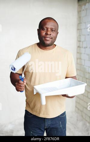 Glücklicher Mann afrikanischer Herkunft mit Paintroller und weißem Quadrat Kunststoffbehälter mit Farbe, die an zwei Wänden in einer Ecke steht Stockfoto