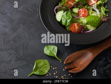 Gesunder, frischer Gemüsesalat mit Salat und Tomaten, roter Zwiebel und Spinat in schwarzer Schüssel auf dunklem Tischhintergrund mit Spatelöffel Stockfoto