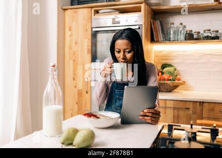 Glückliche schwarze Frau trinkt Kaffee in der Küche zu Hause und mit Tablet. Stockfoto