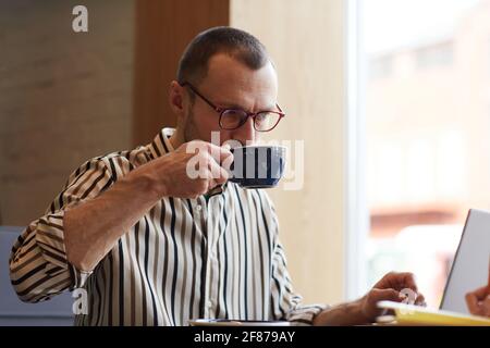 Minimalporträt eines jungen Geschäftsmannes, der Kaffee trinkt, während er mit einem Laptop am Tisch im Café arbeitet, Kopierraum Stockfoto