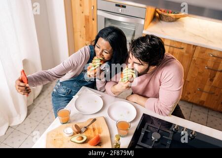 Fröhliche schwarze Frau und ihr Freund haben leckeres Frühstück in der Küche zu Hause und Selfie. Stockfoto