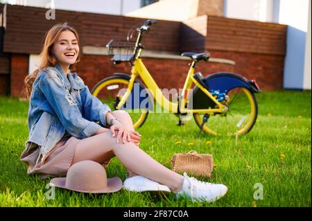 Lächelndes kaukasisches Weibchen (Frau) entspannt sich und sitzt auf dem Gras Im öffentlichen Park vor dem Mietfahrrad Stockfoto