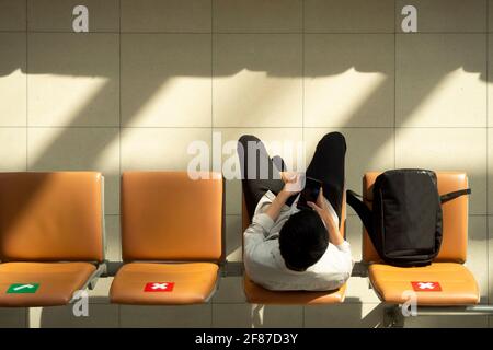 asiatische Geschäftsmann Passagier trägt Maske arbeiten auf Handy und Sitzen Sie auf einem weit entfernenden Stuhl, während Sie auf den Flug warten flughafen-Terminal Durin Stockfoto