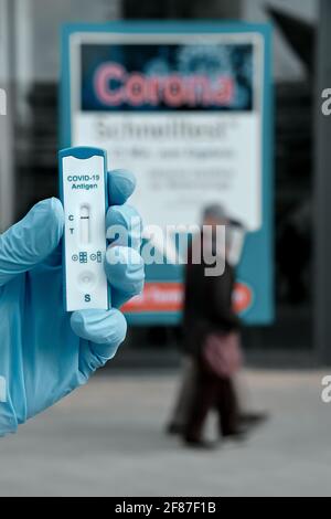 Hand in Handschuh, der den Coronavirus-Test vor dem Banner für Covid-Tests in Mitte hält. Schnelltest, ein schneller Korona-Test in deutscher Sprache, wird für benötigt Stockfoto