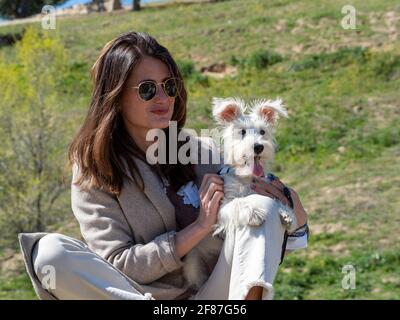 Junge Frau, die auf einem Felsen auf einem Feld sitzt und für ein Foto mit ihrem weißen Schnauzer Welpe posiert.