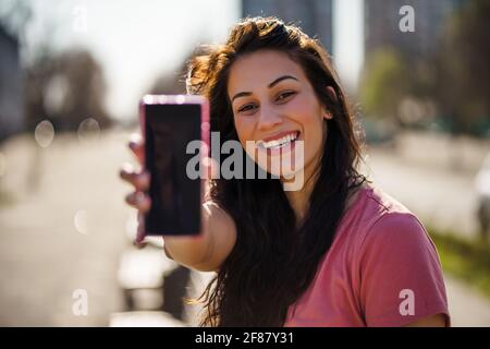 Glückliche junge Frau genießt sonnigen Tag im Freien in der Stadt. Sie hält das Smartphone mit leerem Bildschirm für Ihre Anzeige. Stockfoto