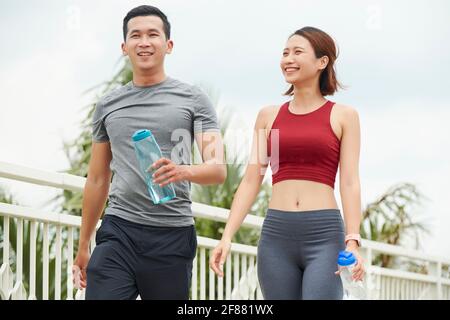Fröhliches, schweißtreibendes junges asiatisches Paar, das im Freien läuft und frisch trinkt Wasser nach dem Training Stockfoto