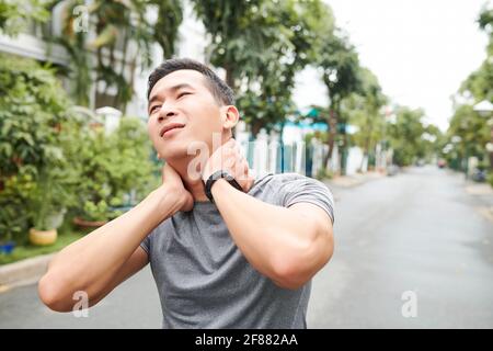 Der junge asiatische Sportler, der unter Schmerzen im Nacken leidet, massiert den Hals, um Erleichterung zu erhalten Stockfoto