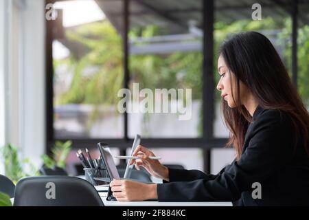 Junge asiatische Frau mit digitalen Tablet mit Stift Stift, während Sitzen an ihrem Schreibtisch im modernen Büro Stockfoto