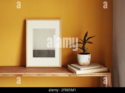 Modell mit Holzrahmen. Blumentopf auf einem Stapel Bücher auf einem alten Holzregal. Moderne Grafiken auf gelbem Hintergrund Stockfoto