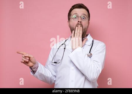 Geschockter Arzt mit einem Stethoskop um den Hals, erschrocken, zeigt den Finger zur Seite, blickt auf die Kamera und bedeckt den Mund mit der Hand. Stockfoto