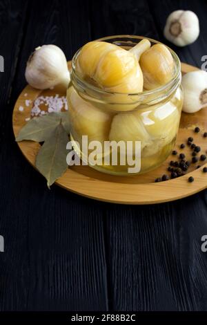 Eingelegter Knoblauch im Glas auf dem braunen Schneidebrett auf dem schwarzen Holzhintergrund.hausgemachtes fermentiertes Produkt. Stockfoto