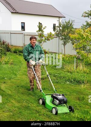 Der Mensch mäht im Garten einen frühlingsgrünen Rasen mit Löwenzahn. Löwenzahn und andere Unkräuter im Hof trimmen. Eine überwucherte Hinterhof-Lichtung mit Pinsel Stockfoto