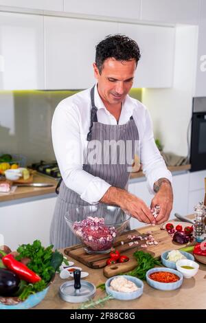 Der Koch fügt den Speck in eine transparente Glasschüssel mit Fleisch und Zwiebeln, während auf der Seite der Arbeitsfläche gibt es Gemüse, Scheiben Speck Stockfoto