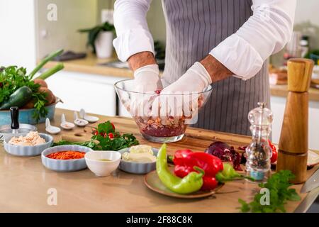 Der Koch Mischen Fleischbällchen mit Gewürzen und Gemüse in einer transparenten Glasschüssel, während auf der Seite der Arbeitsfläche gibt es Gemüse, Scheiben o Stockfoto