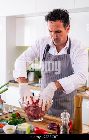 Der Koch Mischen Fleischbällchen mit Gewürzen und Gemüse in einer transparenten Glasschüssel, während auf der Seite der Arbeitsfläche gibt es Gemüse, Scheiben o Stockfoto