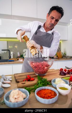 Der Koch fügt die Gewürze in eine transparente Glasschüssel mit Fleisch, Zwiebel und Speck, während auf der Seite der Arbeitsfläche gibt es Gemüse, Scheiben Stockfoto