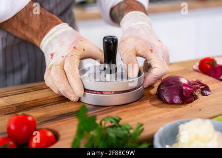 Eine Nahaufnahme eines Küchenchefs, der in einer Presse Fleisch für Burger auf einem Holzbrett in der Küche formt. Es gibt verschiedene Gewürze, Gemüse, sowie kleine Kitk Stockfoto