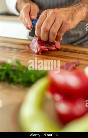 Eine Nahaufnahme der Hände des Küchenchefs, die während der Vorbereitungsphase Carpaccio-Fleisch auf einem Holzbrett in dünne Stücke schneiden. Verschiedene kleine Küche b Stockfoto