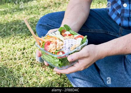 Nahaufnahme, unerkennbarer junger Mann, der auf dem Gras sitzt und einen Glasbehälter mit einem frischen und farbenfrohen Salat hält. Gesunde Lebens- und Ernährungskonzepte. Stockfoto