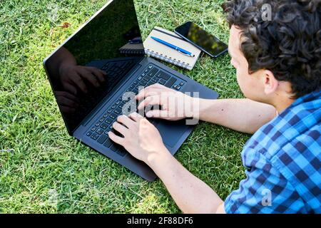 Junger Mann, der einen Laptop im Freien auf dem Rasen benutzt. Konzepte des Fernarbeitens und Studierens. Stockfoto