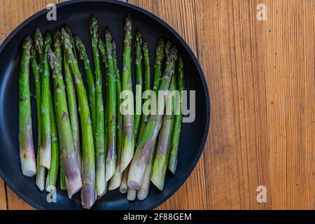 Gebratener grüner Spargel in schwarzer Pfanne auf Holzgrund. Draufsicht, Kopierbereich. Stockfoto