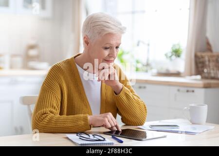 Ältere Geschäftsfrau, die in der Küche sitzt und ein Tablet benutzt Stockfoto