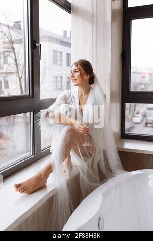 Braut s Morgen. Braut trinkt Champagner im Peignoir. Junge Frau sitzt auf einem großen Fenster in einem Hotelzimmer im Badezimmer. Schönes Mädchen in Stockfoto