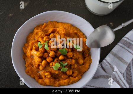 Dicke Kürbissuppe mit gebackenen Kichererbsen und Rettichsprossen auf einem Teller mit einem Glas Milch auf dem Tisch, leckeres gesundes Abendessen Stockfoto