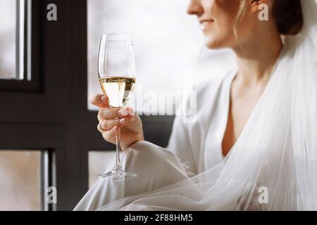 Braut s Morgen. Braut trinkt Champagner im Peignoir. Junge Frau sitzt auf einem großen Fenster in einem Hotelzimmer im Badezimmer. Schönes Mädchen in Stockfoto