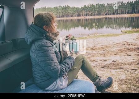 Frau sitzt in ihrem Auto und schaut auf den See, während sie eine warme Mahlzeit aus einer Thermoskanne isst. Das Konzept des Essens draußen und eines Picknicks Stockfoto