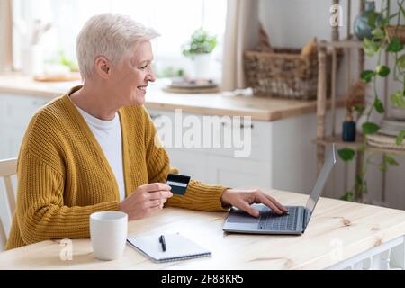 Eine glückliche, ältere Dame, die die Kreditkarte hält und einen Laptop benutzt Stockfoto