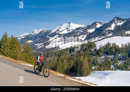 Senior Frau Mountainbiken unter der Nagelfluh-Bergkette mit Hochgrat-Gipfel auf einem E-Mountainbike im Frühfrühling, im Allgäu bei Stei Stockfoto